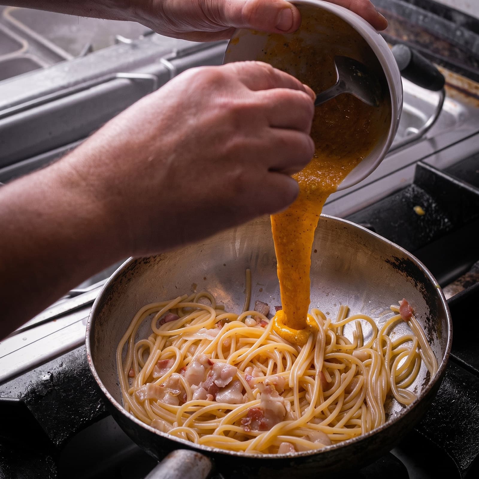 A hand pouring carbonara sauce over spaghetti and pancetta in a pan on a professional stovetop.