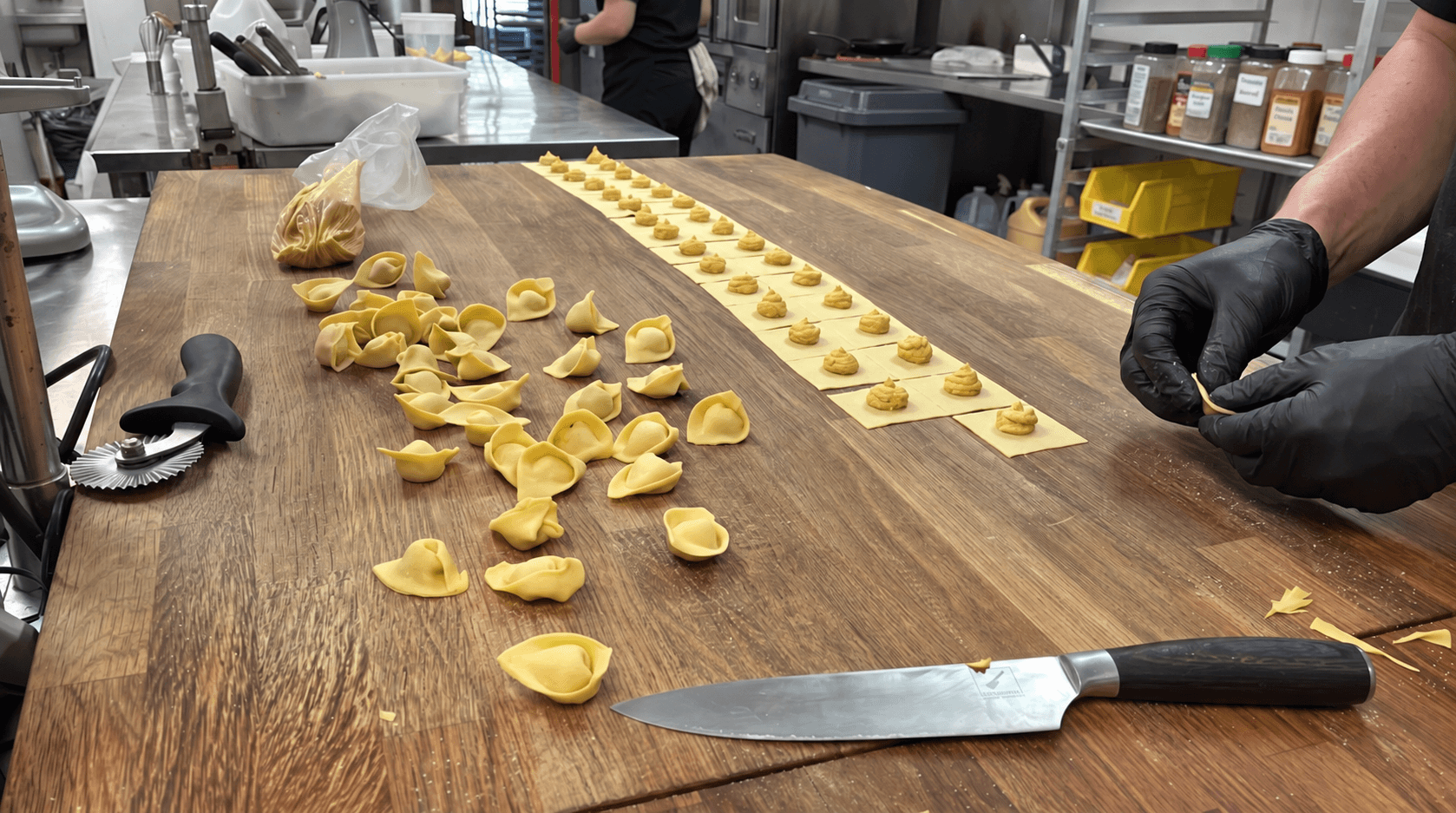 A wood prep table where a gloved hand pinches shut a row of small filled pasta shapes on a pasta sheet, beside a pile of finished pieces, a pastry cutter and a chef's knife, in a professional kitchen.