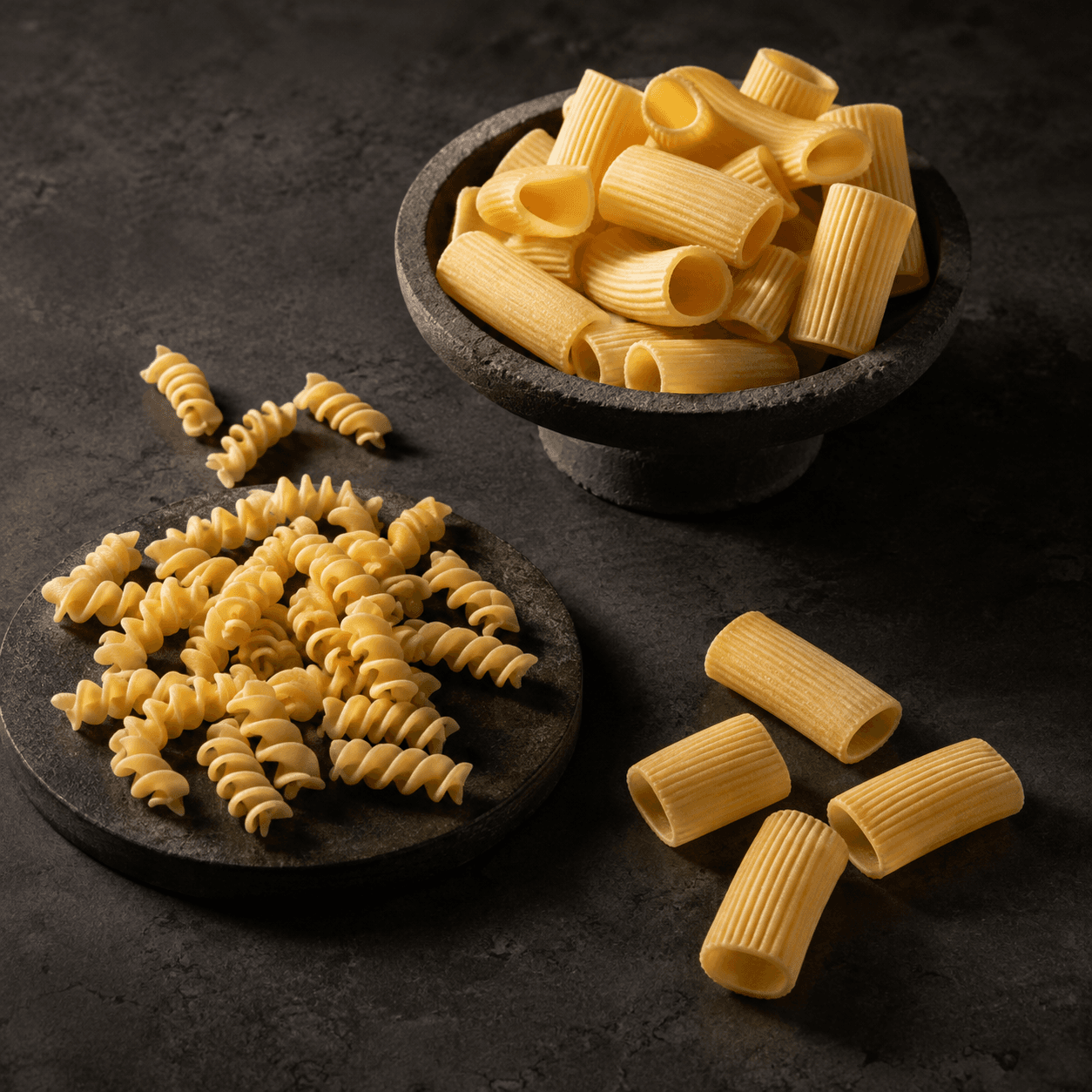Rigatoni in a dark stone bowl and Fusilli on a stone plate, arranged on a dark surface.